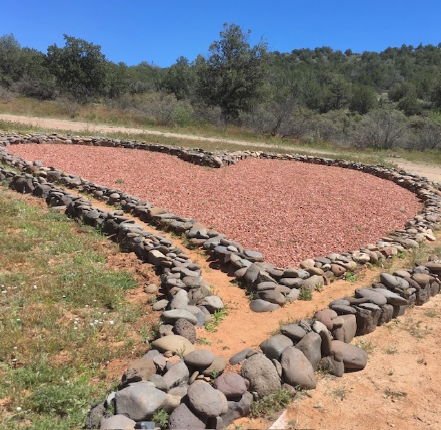 A heart-shaped area filled with red stones is surrounded by a border of smooth rocks, situated in a grassy landscape with trees in the background.